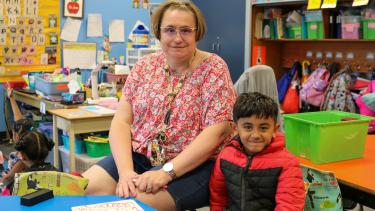 Woman with short hair, wearing glasses and floral shirt sits next to young kindergarten boy in a classroom