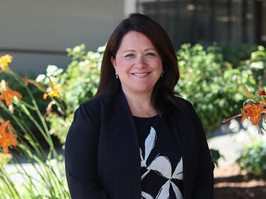 Woman smiles in front of district building and some greenery/flowers in the garden bed