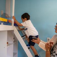 A little boy climbs a ladder to go up to a soft play area while two adults watch and help him get up the ladder.