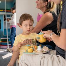 Little boy and his mother playing with a wooden tea set that sits on her lap.
